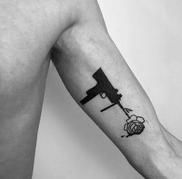 Minimalist black Uzi tattoo on the inner arm, featuring a silhouette design with a rose emerging from the barrel, symbolizing contrast and peace.