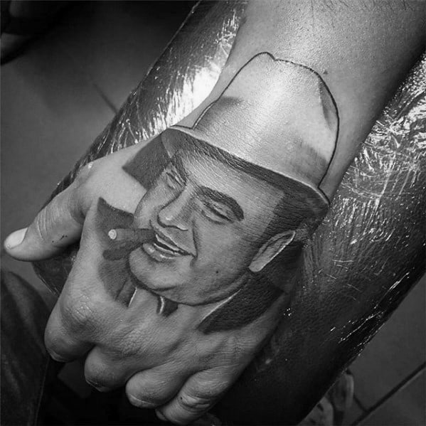 Al Capone tattoo on the hand, featuring a black-and-white portrait of the gangster with a cigar, wearing his signature fedora and smiling.