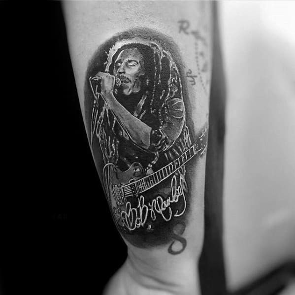 Bob Marley tattoo of him singing and playing guitar, with his signature below, done in black and gray realism on the upper arm.
