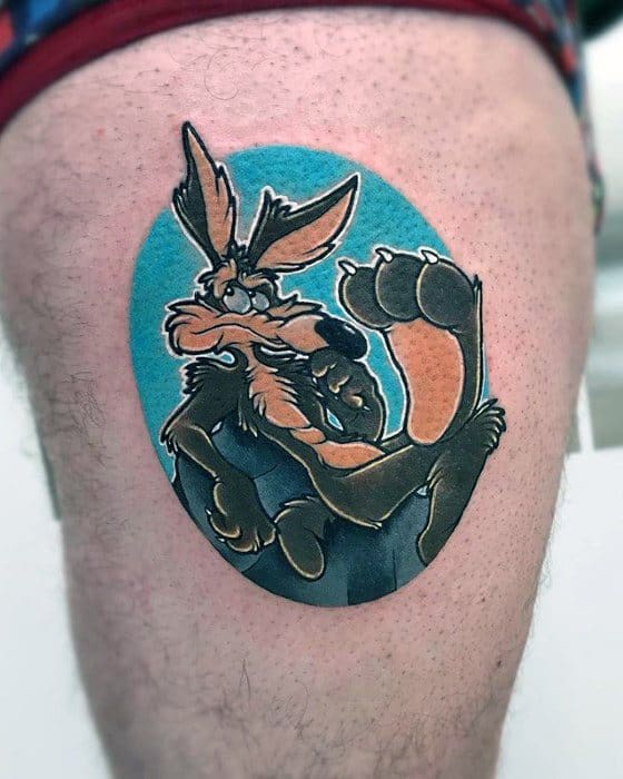 Wile E. Coyote tattoo in a sly, scheming pose with a blue oval background and bold cartoon-style shading on the thigh.