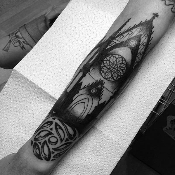 Forearm tattoo of a gothic cathedral with pointed arches, a large rose window, and bold black shading, paired with a Celtic-style pattern.