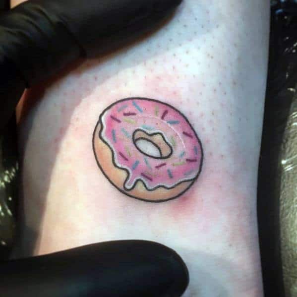 Small tattoo of a pink frosted donut with sprinkles, outlined in black with a simple and cute minimalistic style.