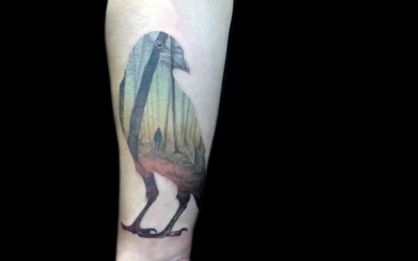 Tattoo of a bird silhouette filled with a forest path and a lone figure walking through tall trees, blending nature and solitude in soft colors.