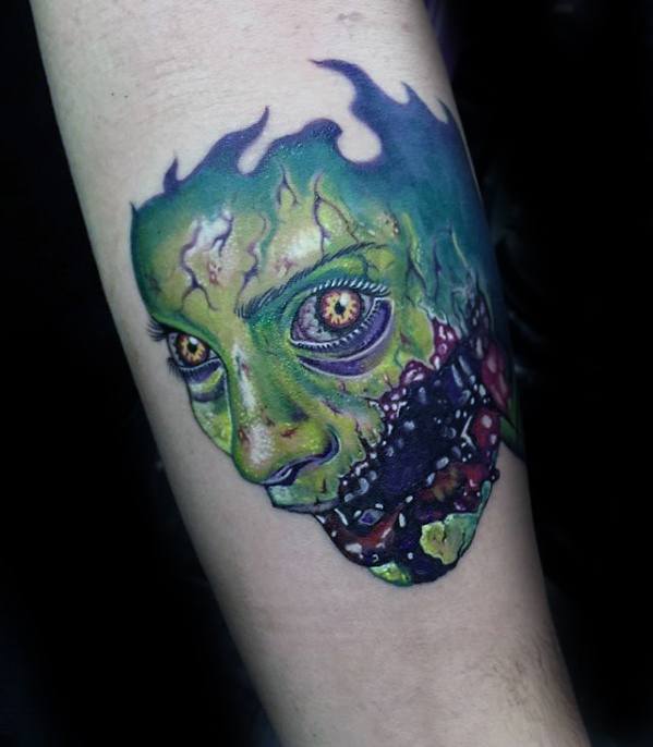 Vibrant Evil Dead tattoo of a possessed face with glowing eyes, green decaying skin, and exposed bloody jaw, framed by a flame-like silhouette