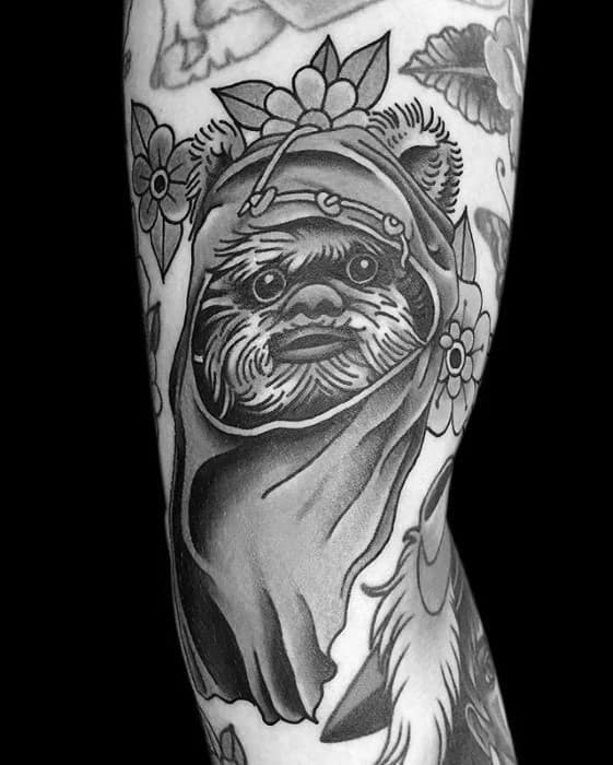 Black and grey Ewok tattoo with a detailed hood and expressive face, surrounded by traditional flower elements in a bold illustrative style.