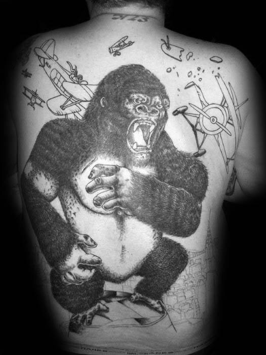 Black and grey back tattoo of King Kong atop a city, gripping a woman and roaring as planes attack, with sketch-style background elements.