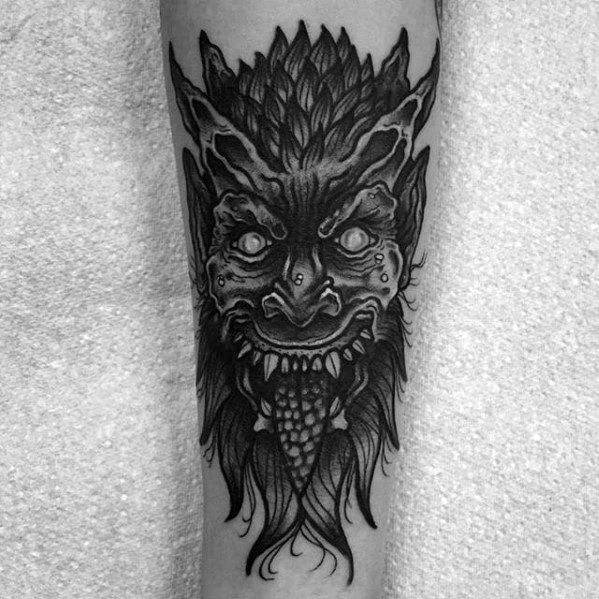 Dark Krampus tattoo in black and grey featuring glowing eyes, textured horns and tongue, sharp teeth, and a sinister expression.