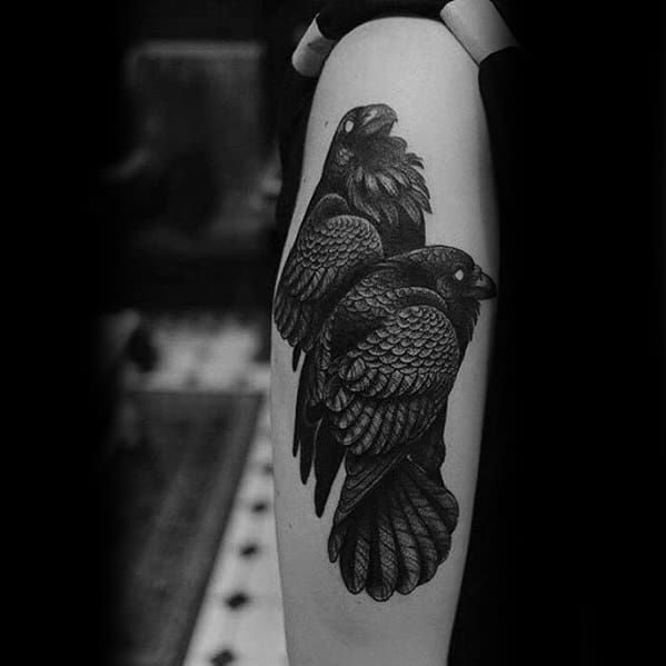 Blackwork tattoo of Huginn and Muninn perched closely together on the upper arm, detailed with layered feathers and bold shading.