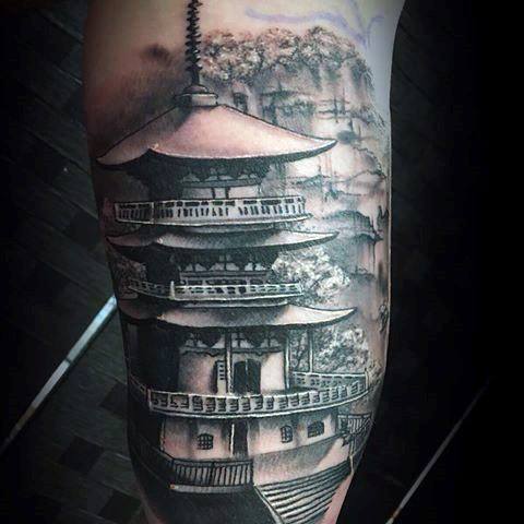 Black and grey tattoo of a multi-level Japanese pagoda with soft background trees and mist, placed on the upper arm.