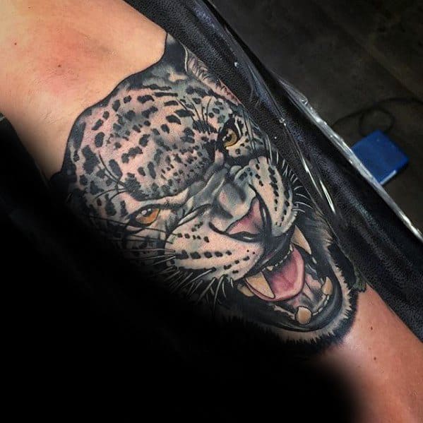 Realistic snow leopard tattoo on the inner arm, showing a snarling expression with lifelike fur texture and piercing amber eyes.