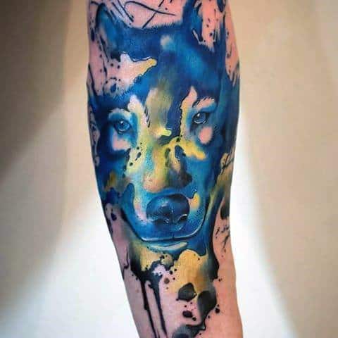 Vivid watercolor wolf tattoo with intense blue and yellow tones, ink splashes, and a bold gaze for a striking forearm design.