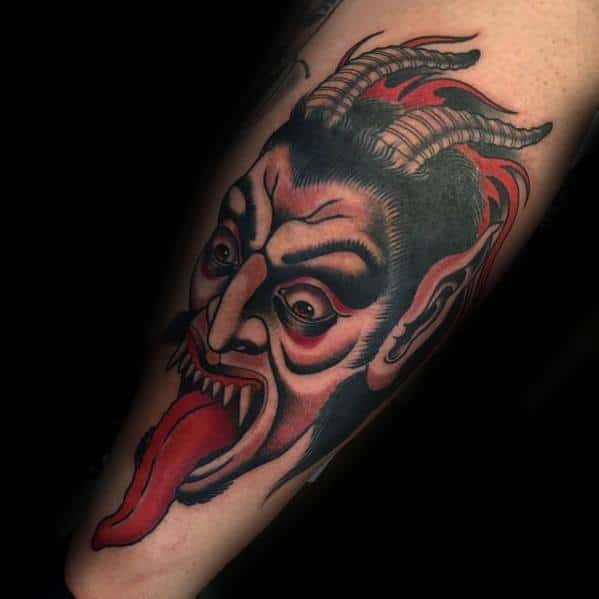Bold Krampus tattoo with a wide-eyed, horned demon face, exaggerated features, and a long red tongue extending dramatically downward.