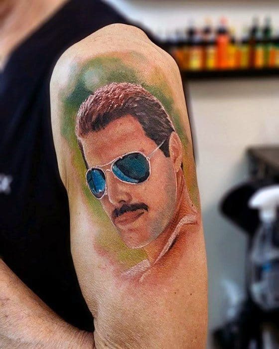 Realistic color tattoo of Freddie Mercury wearing reflective blue sunglasses, with soft background tones and smooth shading on the upper arm.