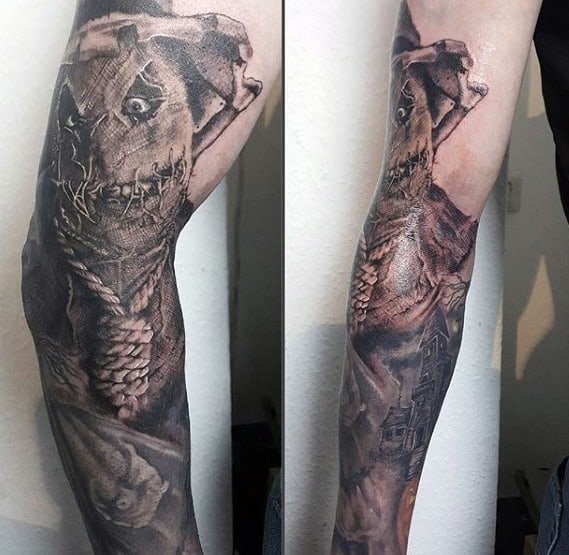 Full sleeve scarecrow tattoo with a sinister stitched face, rope collar, eerie shadows, and a haunted house in detailed black and grey ink.