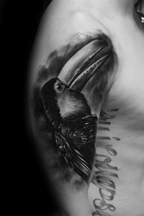Realistic black and grey toucan tattoo on the upper arm with soft shading and a dramatic cloudy background for a moody effect.