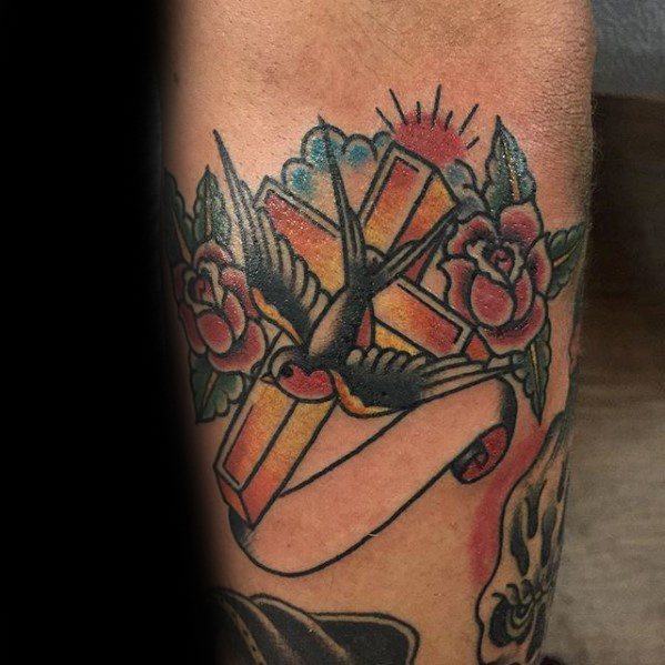 Traditional-style tattoo of a 3D cross with a swallow, red roses, and sun rays, featuring a blank ribbon wrapped around the base.