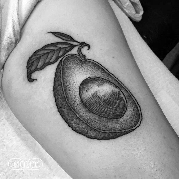 Black and grey avocado tattoo with a detailed, textured shell pit and leaf stem, done in fine line and stippling style on the upper arm.