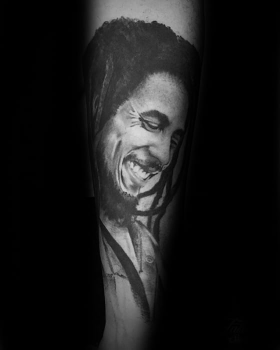 Realistic black and gray tattoo of Bob Marley with eyes closed and a gentle smile, detailed dreadlocks flowing over the shoulder.
