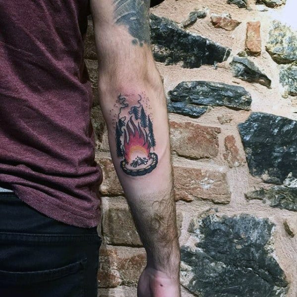 Forearm tattoo of a small campfire with glowing red and orange flames, smoky accents, and a stone ring in a stylized illustrative design.