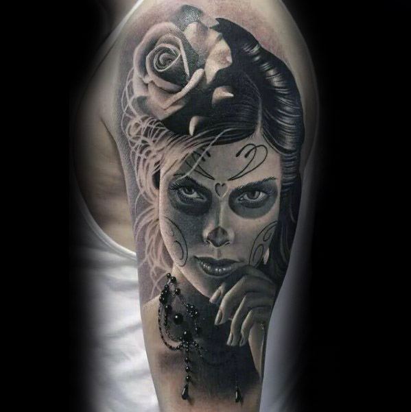 La Catrina tattoo of a woman with intense gaze, sugar skull makeup, a rose in her hair, and a beaded necklace on her hand.