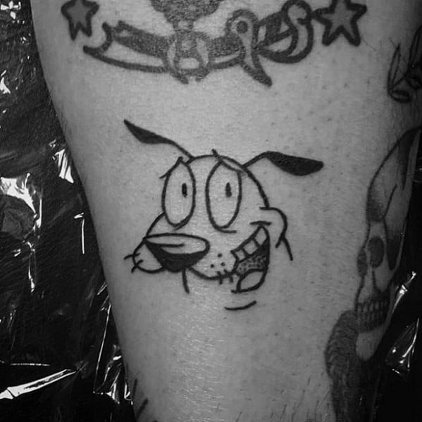 Small black ink tattoo of Courage the Cowardly Dog’s face with a goofy, slightly nervous smile, placed on the lower leg.