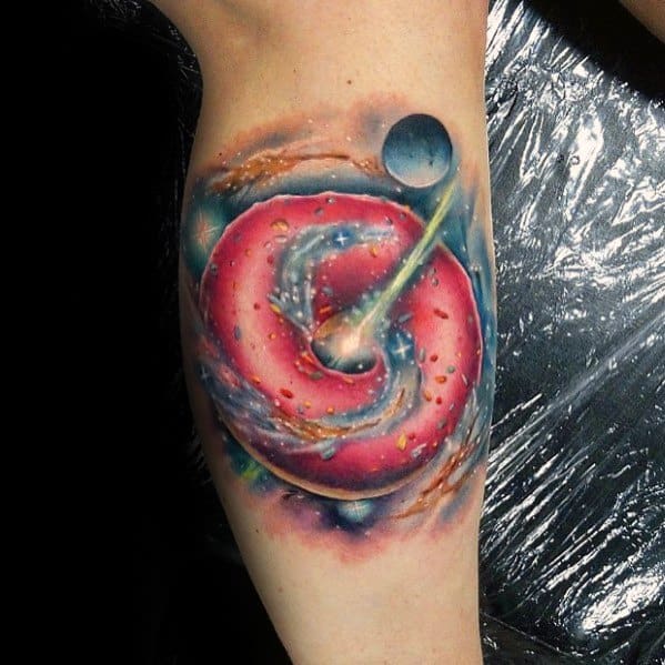 Tattoo of a pink frosted donut swirling in a galaxy with planets and cosmic elements, blending space and sweets in a surreal design.