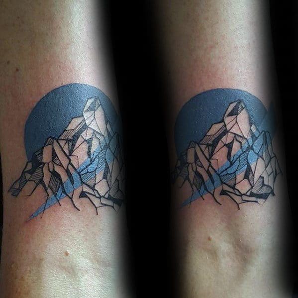 Geometric mountain tattoo with sharp black outlines, accented by a bold blue circle and shaded blue glacier streaks.