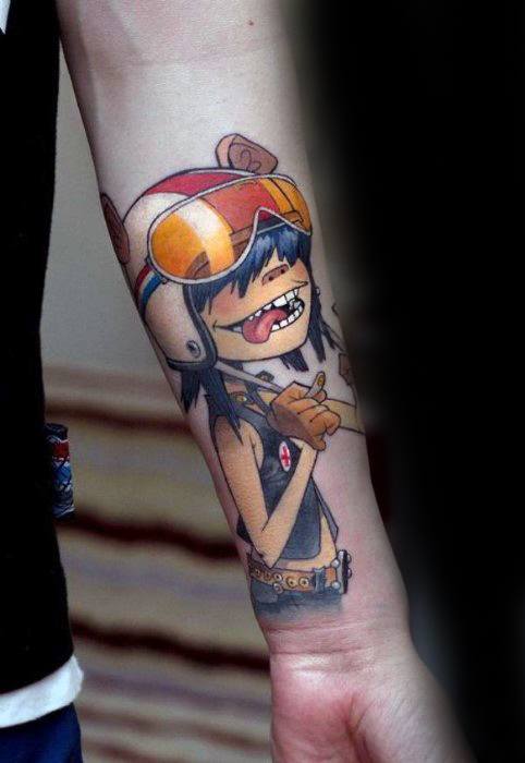 Colorful forearm tattoo of Noodle from Gorillaz wearing a bear-eared helmet and orange visor, smiling confidently in a rocker outfit.