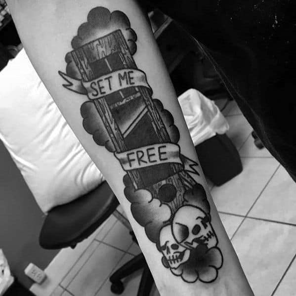 Black and grey guillotine tattoo surrounded by clouds, two skulls at the base, and a banner wrapping around with the words “SET ME FREE.”