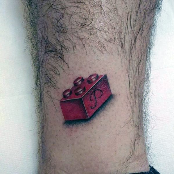 Small tattoo of a red Lego brick with a cursive letter “P” on the side, featuring realistic shading and a slight 3D effect on the leg.