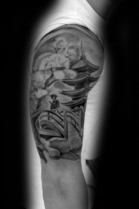 Black and grey upper arm tattoo of a Japanese pagoda with a cherry blossom tree, arched bridge, and a figure crossing under cloudy skies.