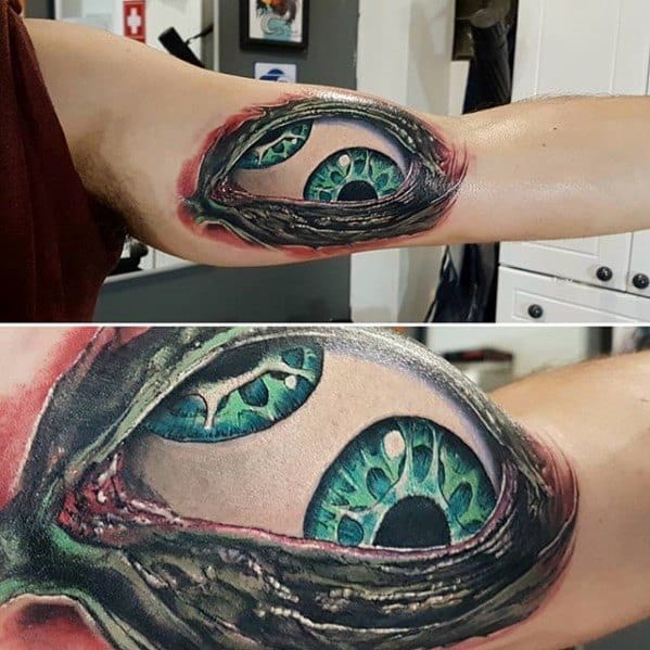 Tool-inspired inner arm tattoo with two vivid green-blue eyes revealed through a torn skin illusion, surrounded by detailed shading and redness.