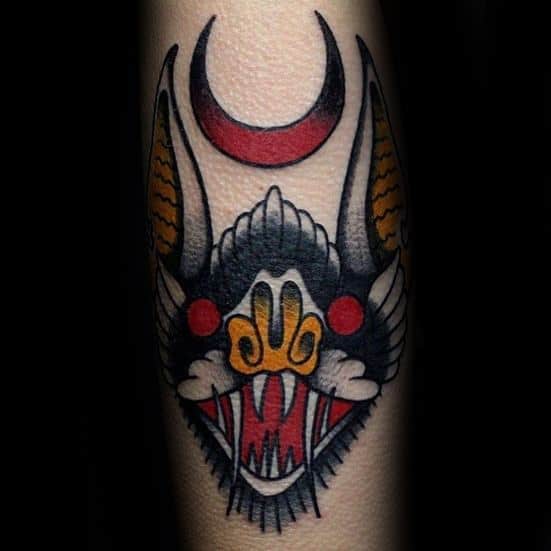 Traditional bat head tattoo with red crescent moon, sharp fangs, and bold black, yellow, and red details for a fierce look.