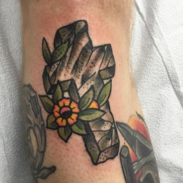 Stone-textured cross tattoo with a yellow and orange flower and green leaves, designed in a bold traditional style on the leg.