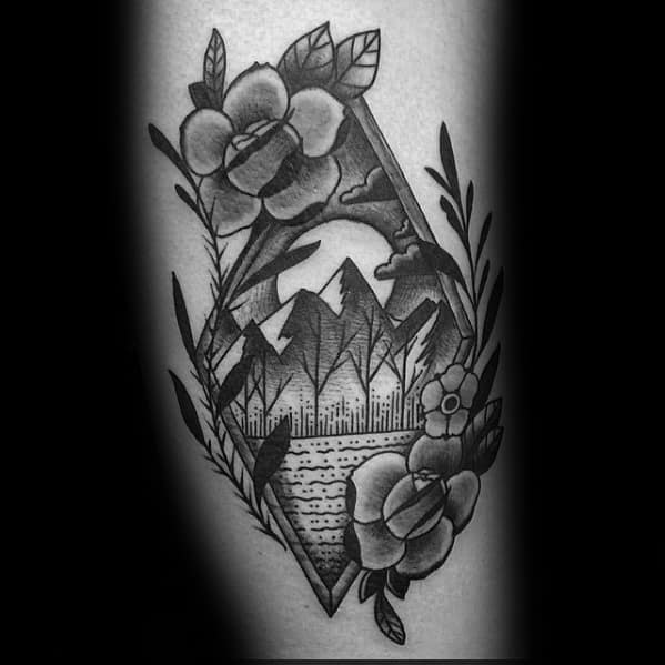 Black and grey diamond-shaped tattoo with a forest, mountains, moon, and water, framed by detailed flowers and leafy branches.