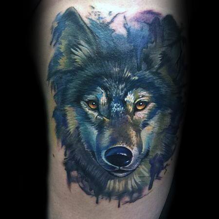 Realistic wolf tattoo with piercing amber eyes, detailed fur texture, and subtle watercolor accents in blue and purple hues.