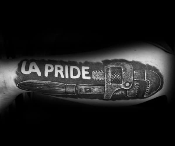 Bold black and grey tattoo of a large pipe wrench with the words LA PRIDE carved out in negative space above it on the inner arm.