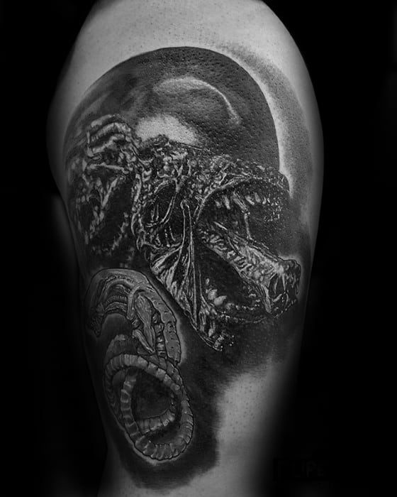 Black and grey Xenomorph tattoo on upper arm showing a roaring alien with detailed textures and a curled tail design below.