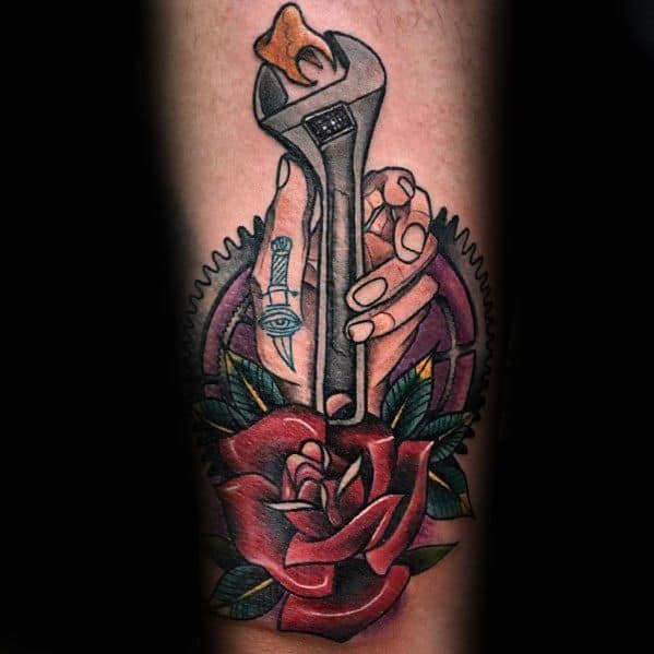 Color tattoo of a hand holding a wrench with a flame at the top, surrounded by a red rose, gear, and an eye-in-hand symbol on the wrist.