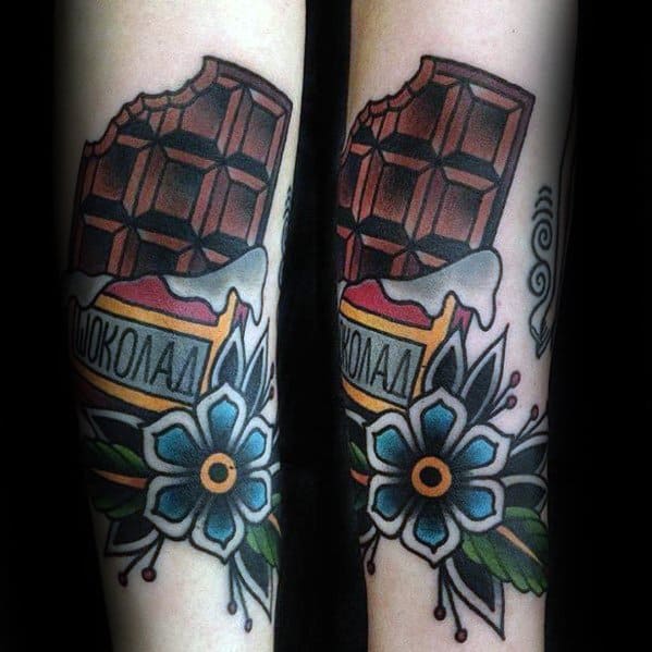 Colorful tattoo of a bitten chocolate bar with a wrapper featuring Cyrillic text, surrounded by blue flowers and bold traditional-style shading.