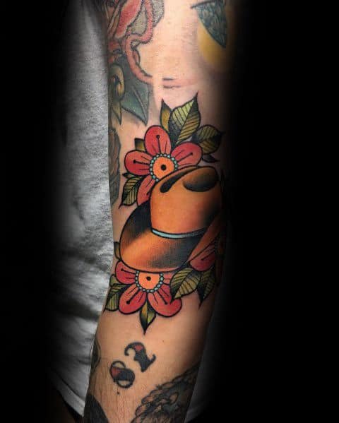 Tattoo of a cowboy hat surrounded by red flowers and green leaves, done in a bold traditional style with strong outlines and vibrant colors.