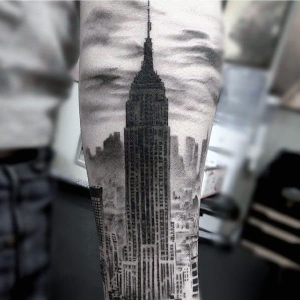 Realistic Empire State Building tattoo featuring a detailed cityscape and cloudy sky, inked on a man's forearm in rich black shading.