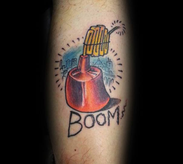 Tattoo of a lit dynamite plunger with "BOOM" written below, featuring bold shading, a cityscape background, and a comic-style explosion effect.