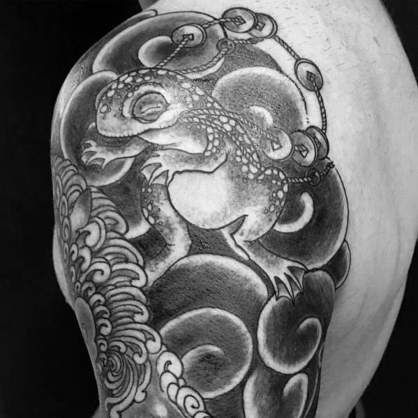 Black and grey Japanese-style shoulder tattoo featuring a mystical toad with gold coins, surrounded by swirling clouds and intricate wave patterns for a traditional look.