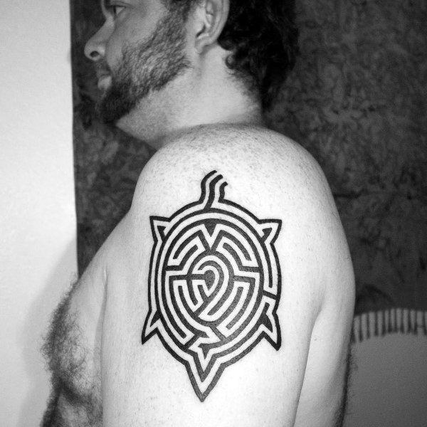 Bold black ink turtle-shaped labyrinth tattoo on a man's upper arm, featuring an intricate maze design within the turtle's shell outline.