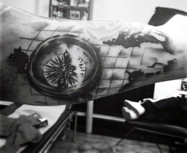Black and grey tattoo of a detailed compass overlaid on a world map grid, creating a vintage exploration theme on the inner bicep