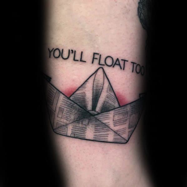 Paper boat tattoo with 'YOU'LL FLOAT TOO' text, inspired by IT, symbolizing eerie nostalgia and a connection to the horror classic.