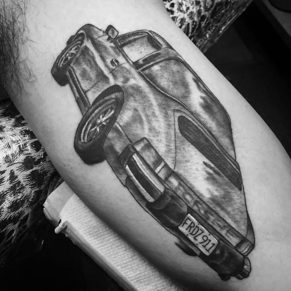 Bold, angled Porsche car tattoo in detailed black shading, featuring distinctive license plate on the forearm.