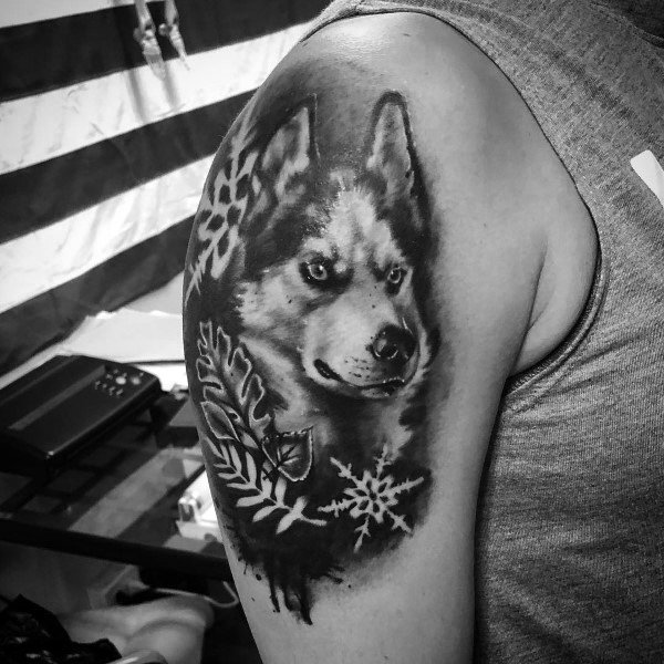 Black and gray Siberian Husky tattoo on a man's upper arm, featuring snowflakes, leaves, and a detailed fur texture for a winter-inspired design.
