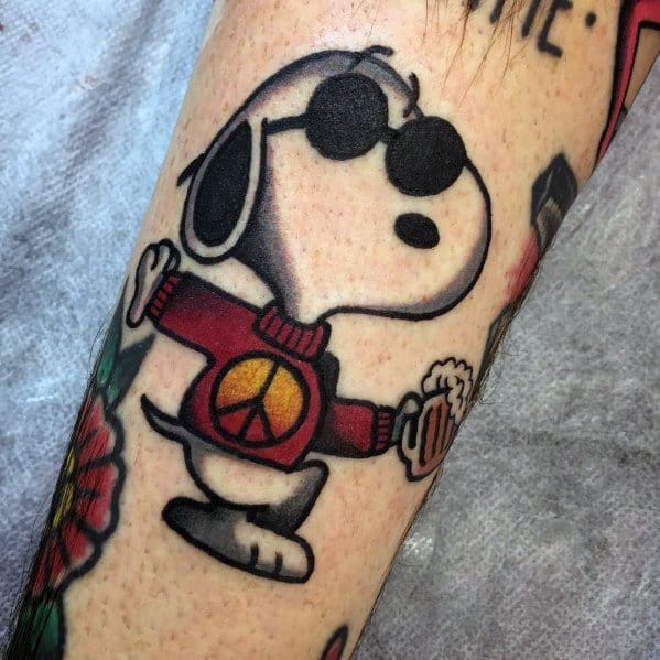 Colorful tattoo of Snoopy wearing sunglasses and a red sweater with a peace symbol, holding a beer mug, placed on a person's arm.